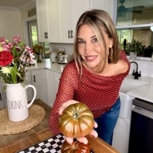 A woman in a red polka dot top smiles in a kitchen, holding heirloom tomatoes over a checkered cutting board. Flowers and a pitcher labeled DRINK sit on the counter beside her.
