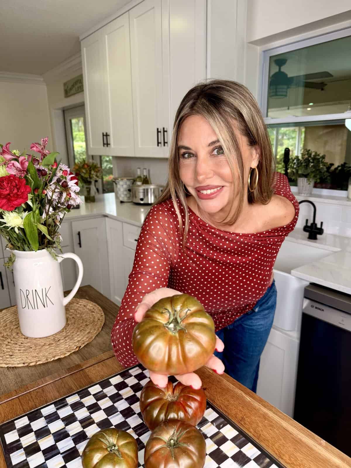 A woman in a red polka dot top smiles in a kitchen, holding heirloom tomatoes over a checkered cutting board. Flowers and a pitcher labeled DRINK sit on the counter beside her.