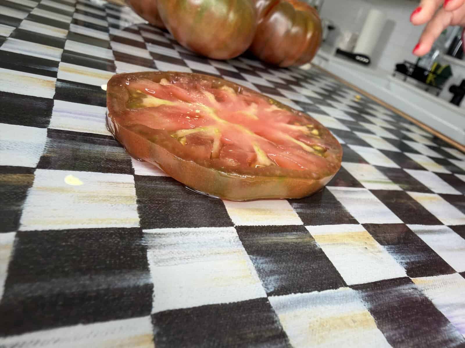 A slice of tomato sits on a black and white checkered surface, with whole tomatoes and a hand visible in the background.