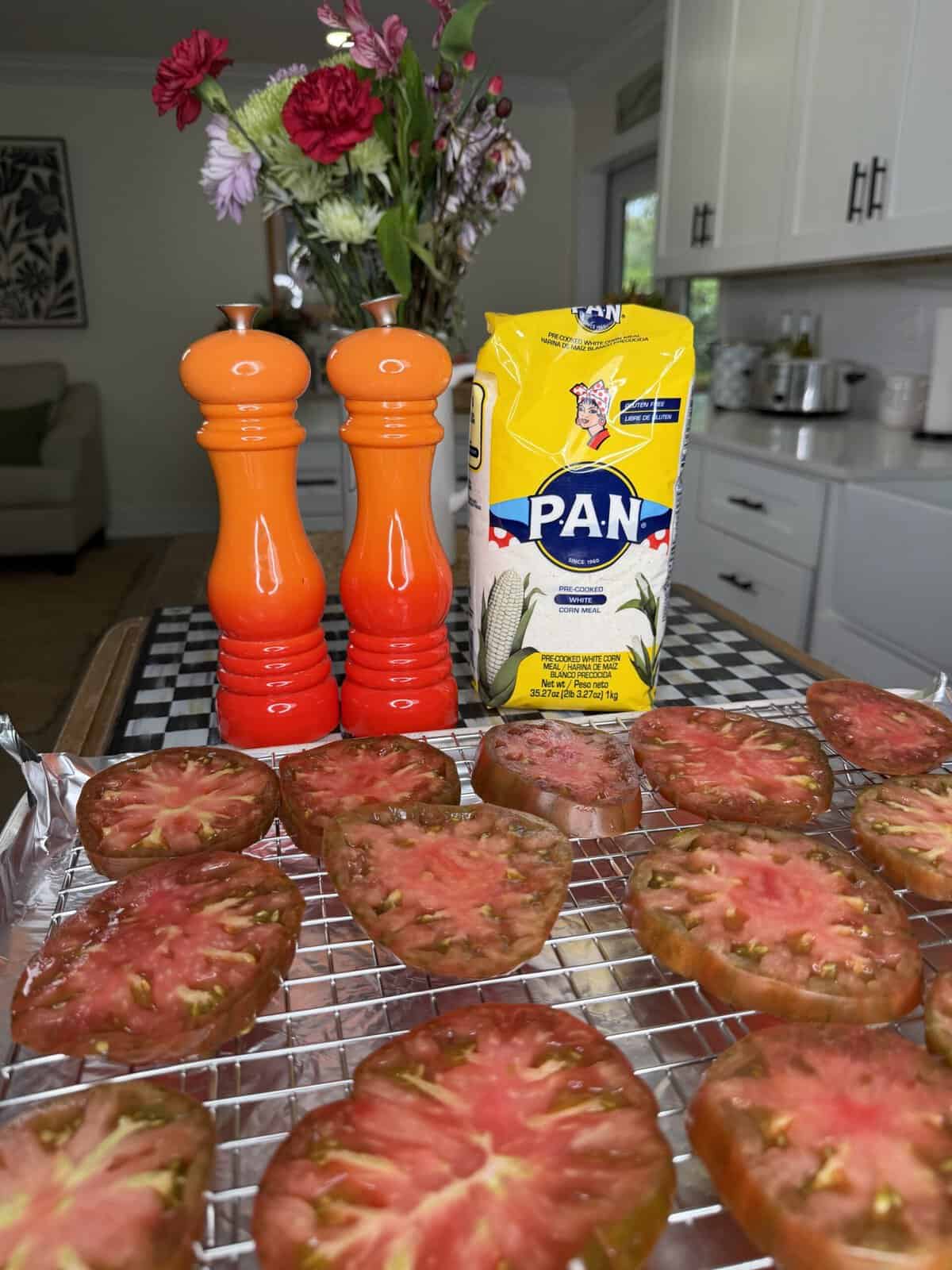 Sliced tomatoes on a cooling rack, two orange shakers, and a bag of PAN cornmeal sit on a kitchen counter. A vase with flowers is in the background.
