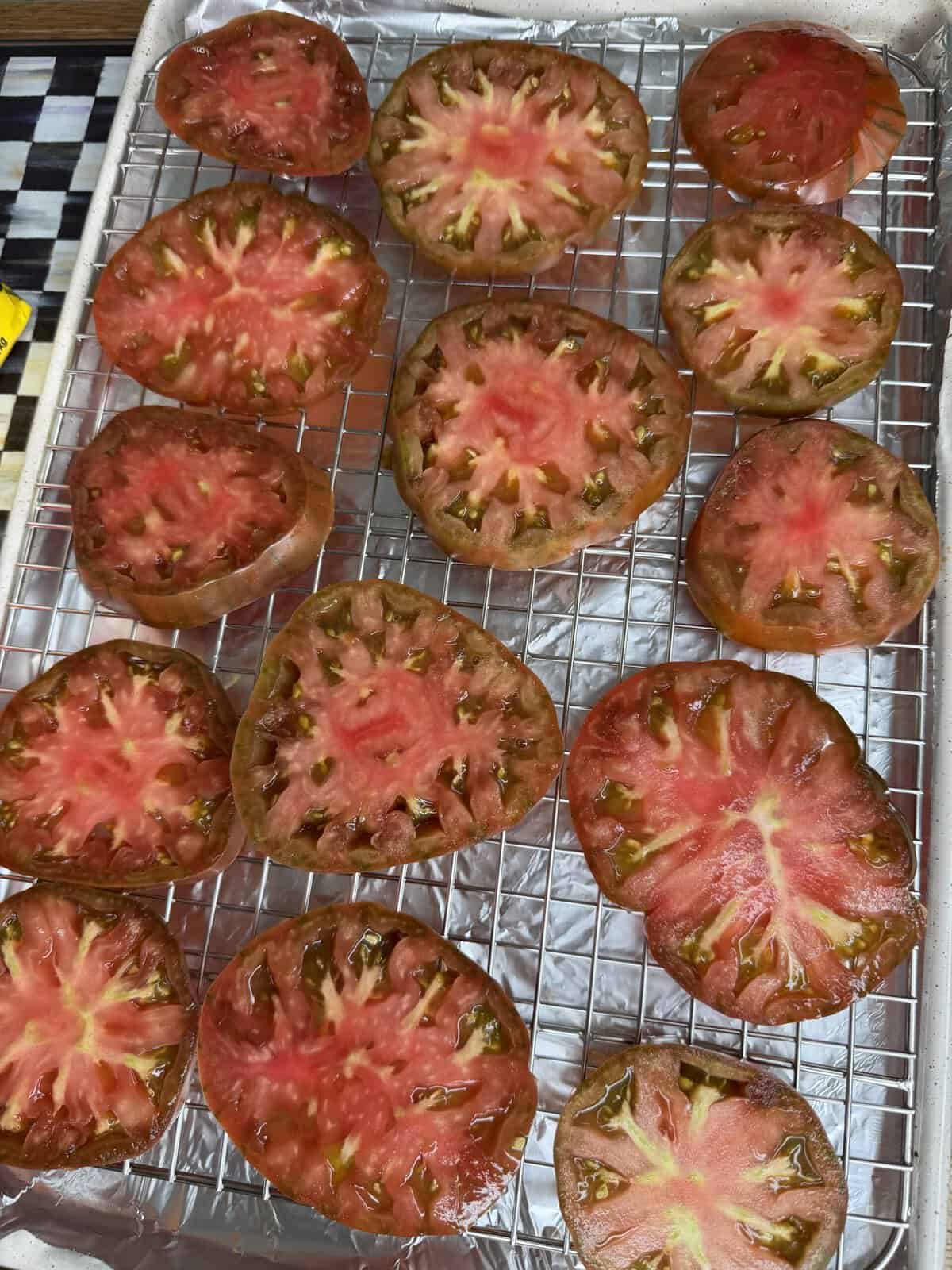 Sliced heirloom tomatoes with red and green hues are arranged on a wire rack over a foil-lined baking sheet. The seeds and inner textures are visible.
