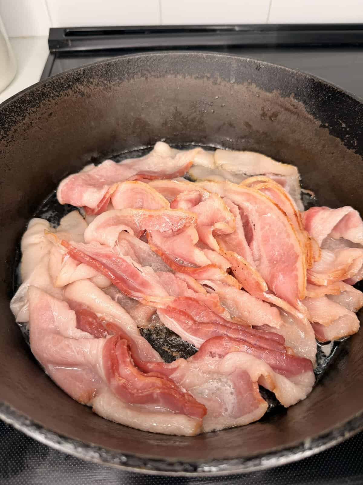Raw bacon strips sizzling in a black cast iron skillet on a stovetop, beginning to cook with some fat starting to render but most pieces still pink and uncooked.