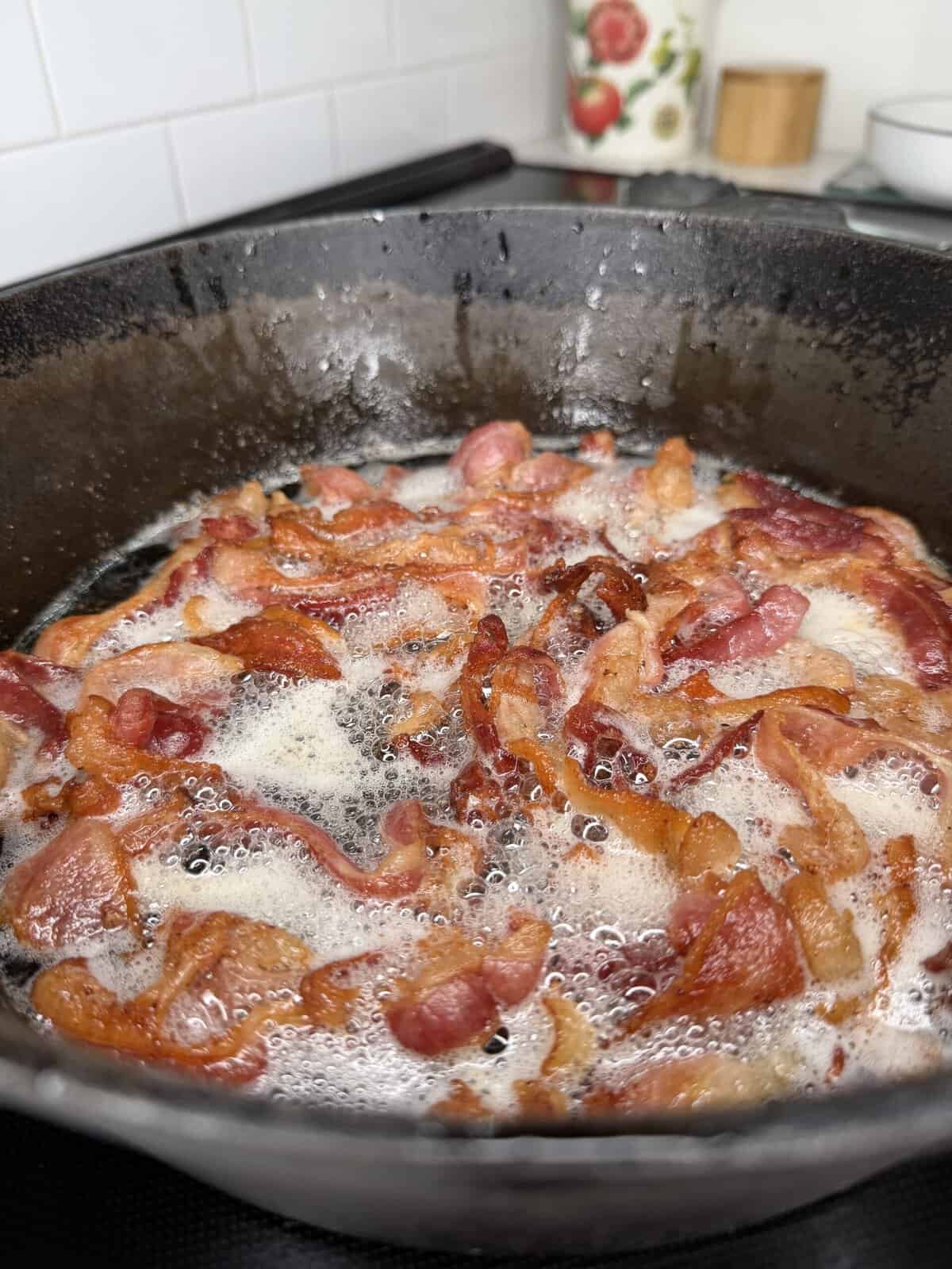 Crispy bacon strips sizzling in bubbling grease in a black cast iron skillet on a stovetop, with a white tiled backsplash in the background.