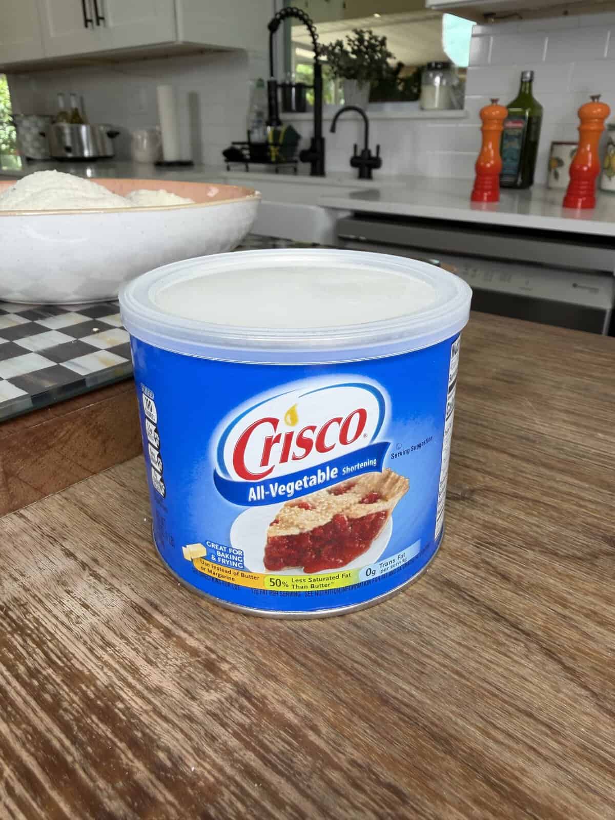 A blue tub of Crisco All-Vegetable Shortening sits on a wooden kitchen counter, with a white bowl of flour and various kitchen items visible in the background.