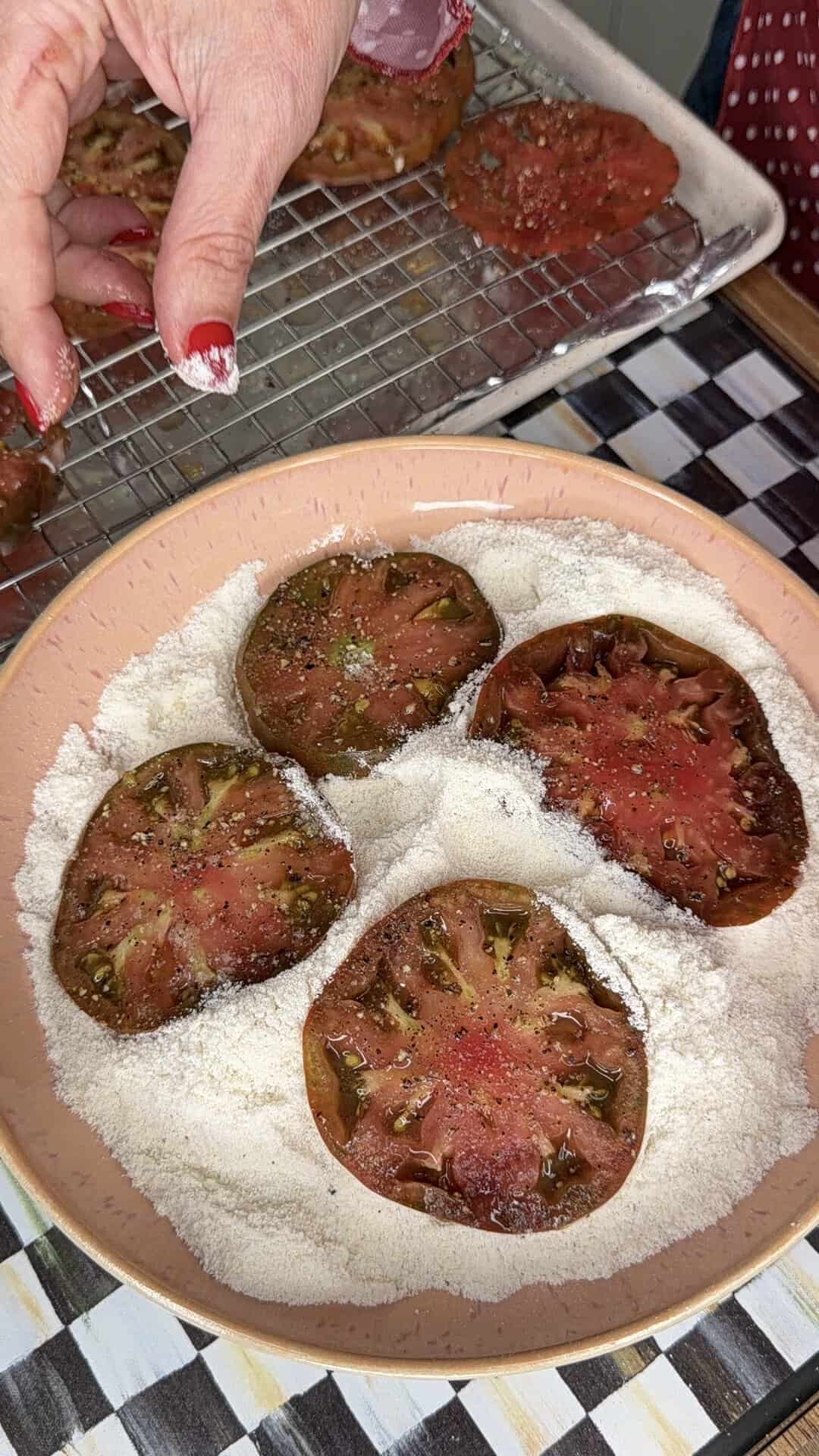 A hand sprinkles flour over thick slices of tomato in a bowl of flour, with more tomato slices on a cooling rack in the background.