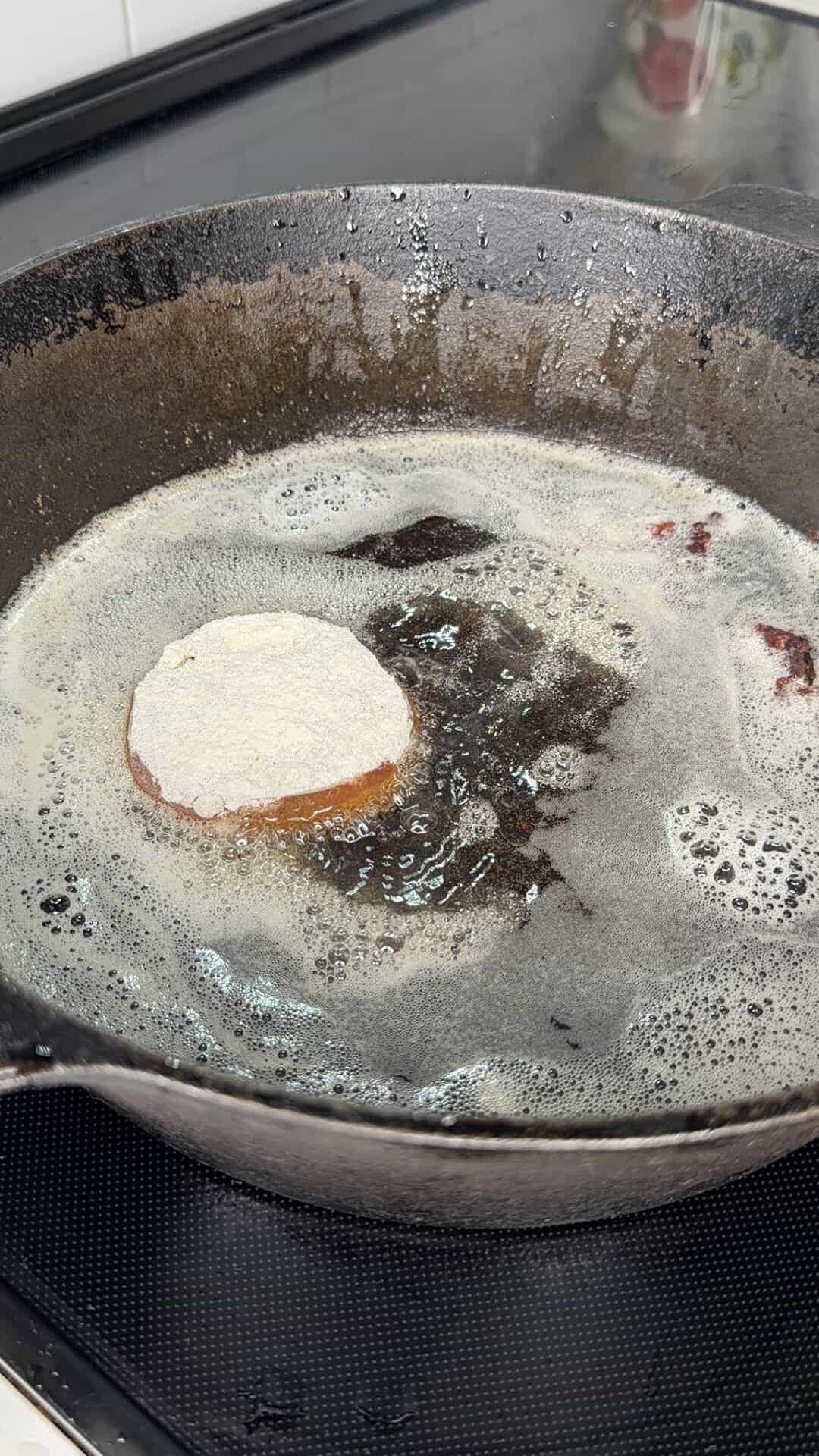 A breaded round slice, possibly a vegetable or meat, is frying in a cast iron pan with bubbling oil on a stovetop.