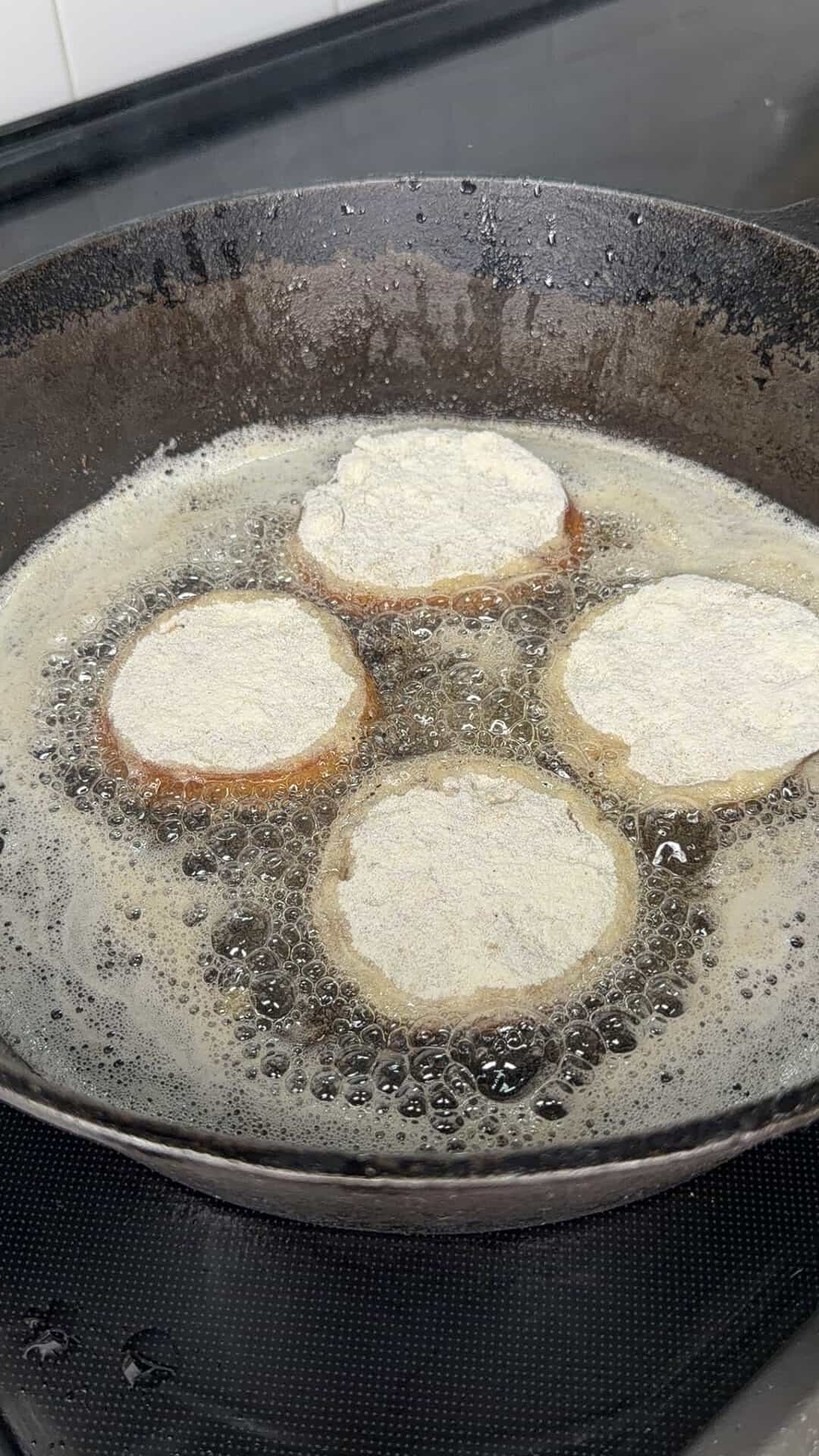 Four floured slices of food are being fried in hot oil in a cast iron pan, with bubbles forming around them. The stovetop and kitchen tiles are visible in the background.