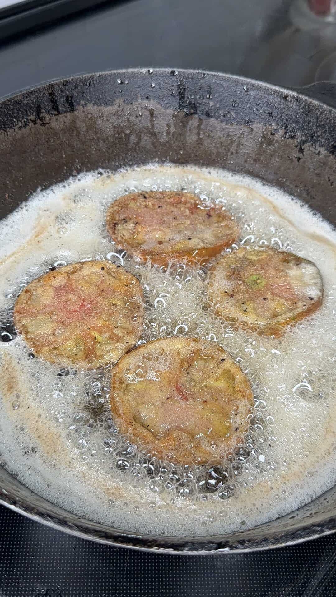 Four battered slices of what appears to be green tomatoes are frying in hot oil in a black skillet, with bubbles forming around them as they cook.