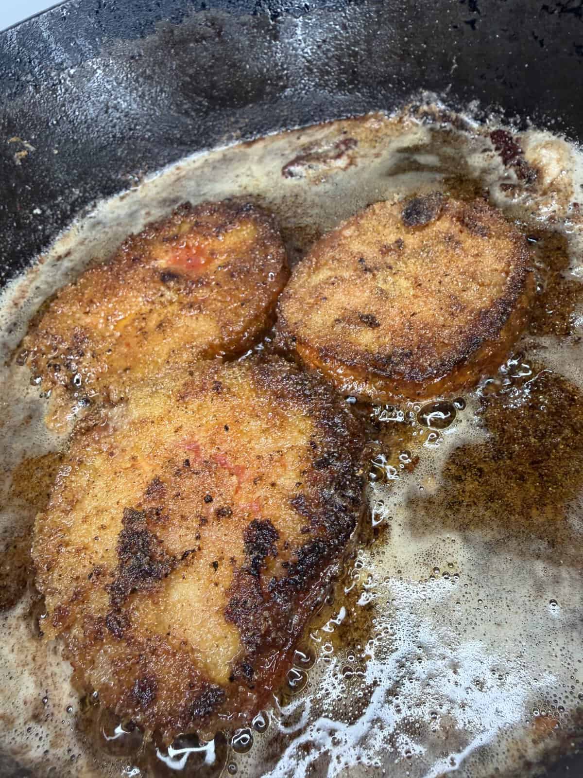 Three breaded and fried slices of food, possibly tomatoes or eggplant, sizzling in hot oil in a pan, with a crispy golden-brown crust.