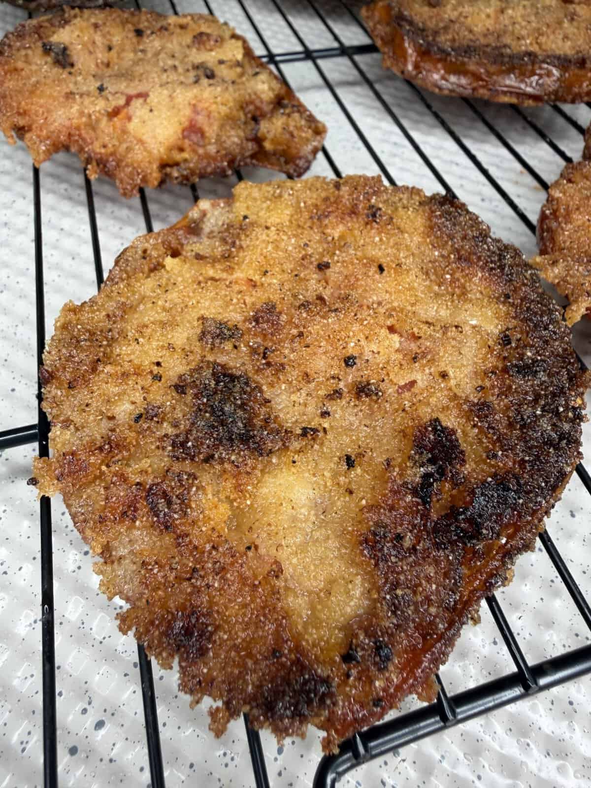 A close-up of a crispy, golden-brown breaded and fried round food item resting on a black wire cooling rack with more pieces in the background.