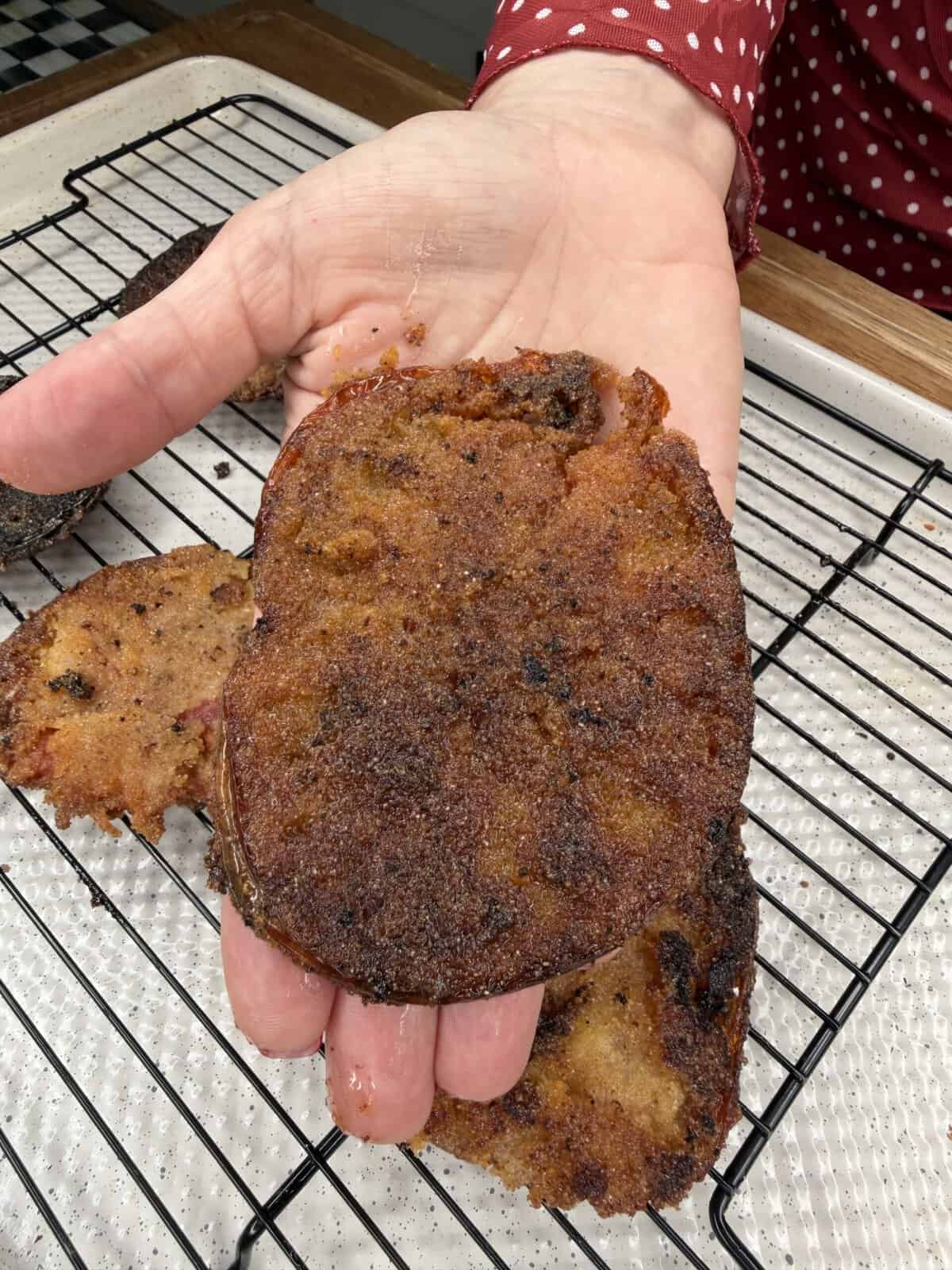 A hand holds up a piece of fried food with a dark, crispy, uneven surface over a cooling rack; another piece rests below it. The person is wearing a red shirt with white polka dots.