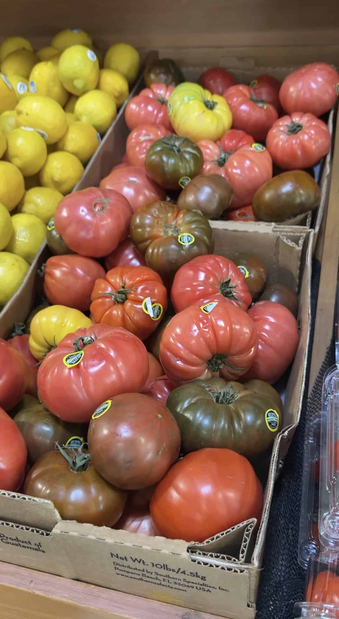 Heirloom tomatoes of various colors and sizes are displayed in a cardboard box next to yellow lemons at a grocery store. A container of grape tomatoes is visible on the right edge.
