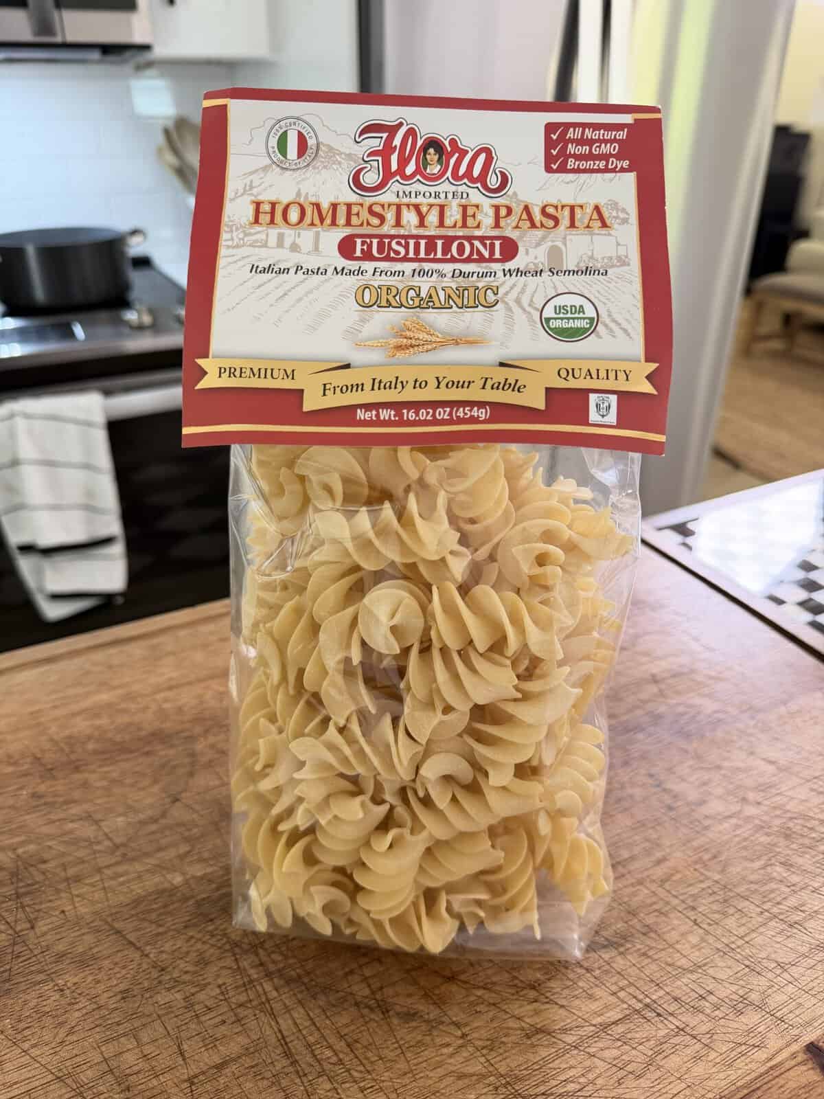 A bag of Flora Homestyle Pasta Fusilloni sits on a wooden counter in a kitchen. The pasta is organic, made from durum wheat, and the packaging is labeled with various quality and organic certifications.