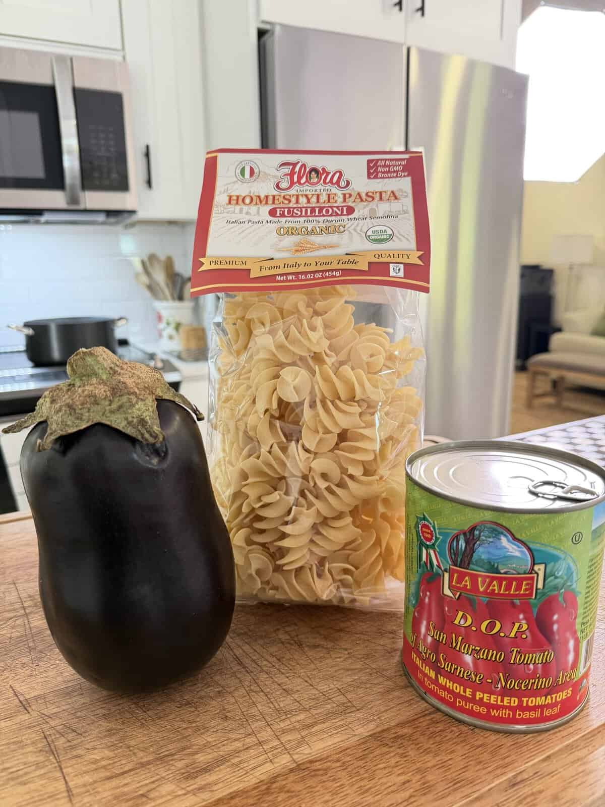 A raw eggplant, a bag of fusilloni pasta, and a can of San Marzano tomatoes are arranged on a wooden kitchen counter with a stove and white cabinets in the background.