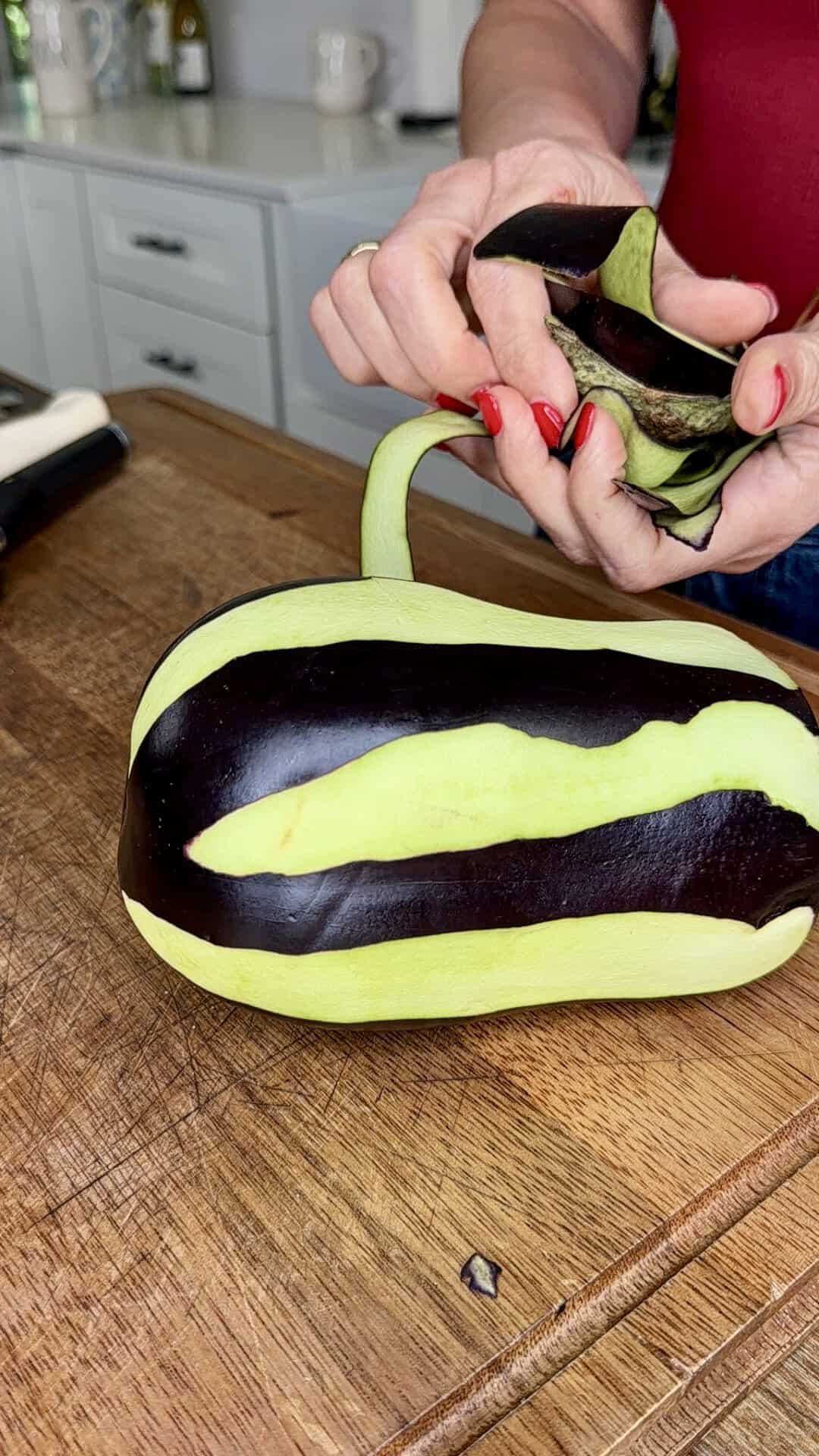 A person with red nails peels stripes of dark purple skin off a light green eggplant on a wooden cutting board in a kitchen.