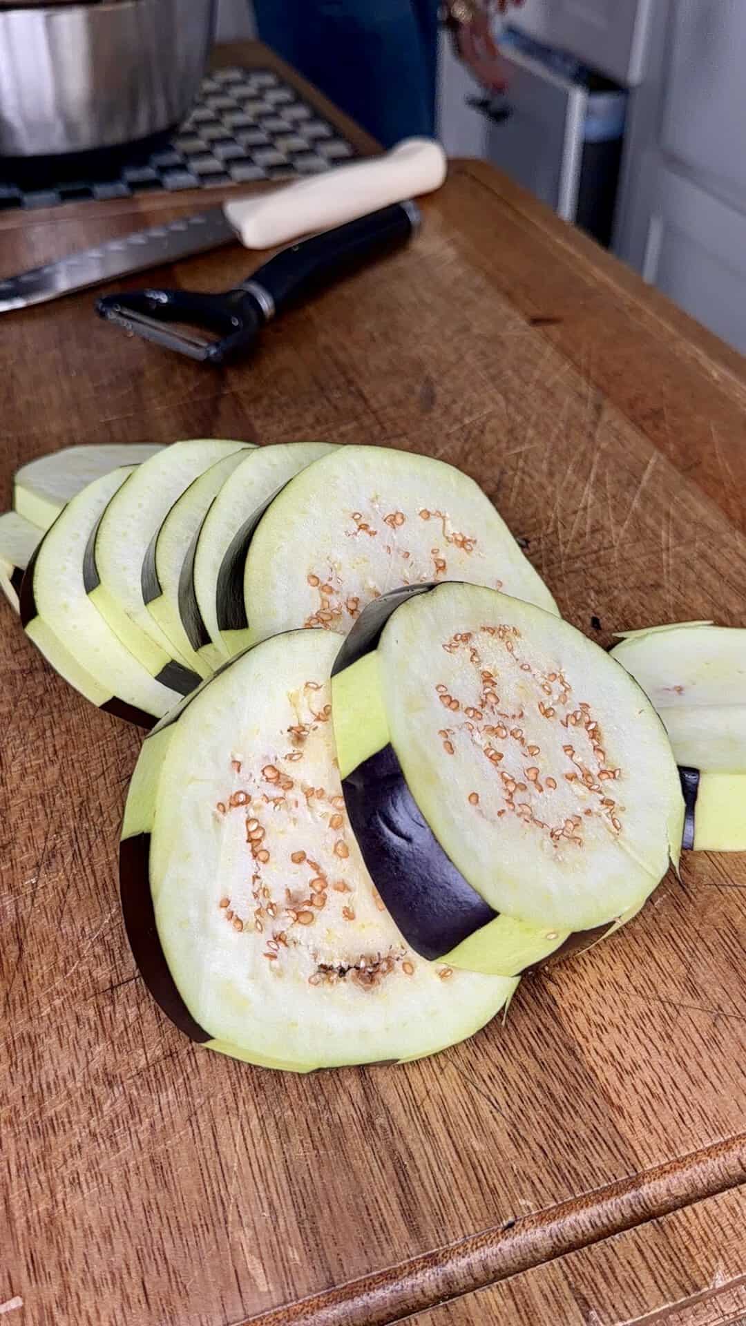 Sliced eggplant rounds are arranged on a wooden cutting board, with a vegetable peeler and knife visible in the background.