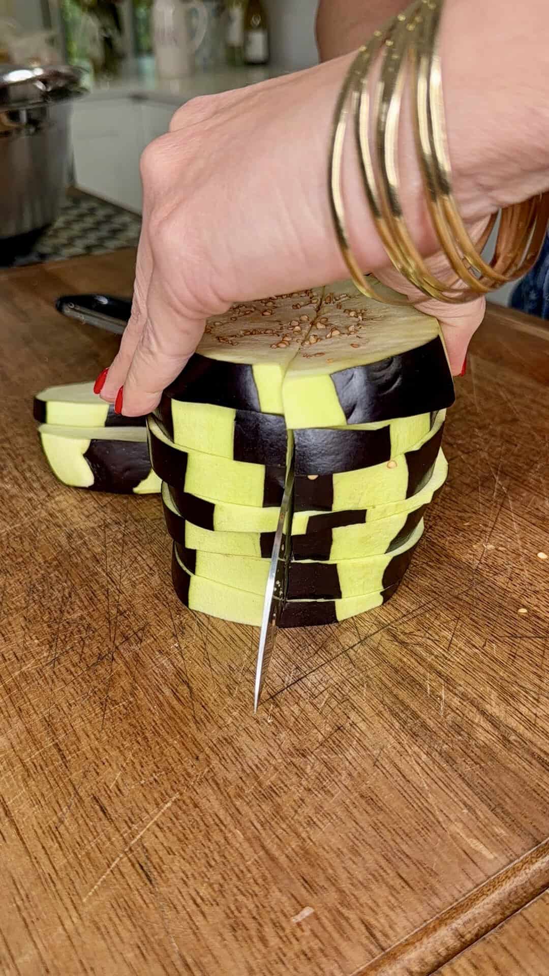 A hand with gold bangles holds a partially peeled eggplant being sliced on a wooden cutting board, showing a checkerboard pattern on the vegetable.