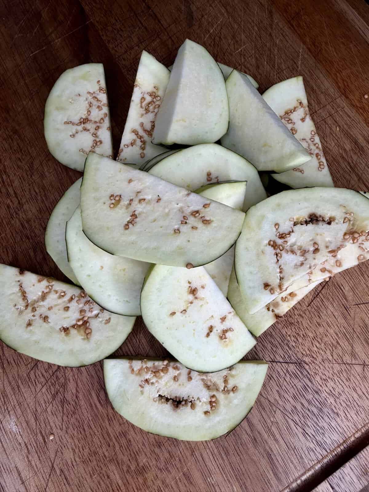 Slices of raw eggplant with visible seeds are arranged on a wooden cutting board. The eggplant pieces are pale with a light greenish hue and some brown seeds scattered throughout.