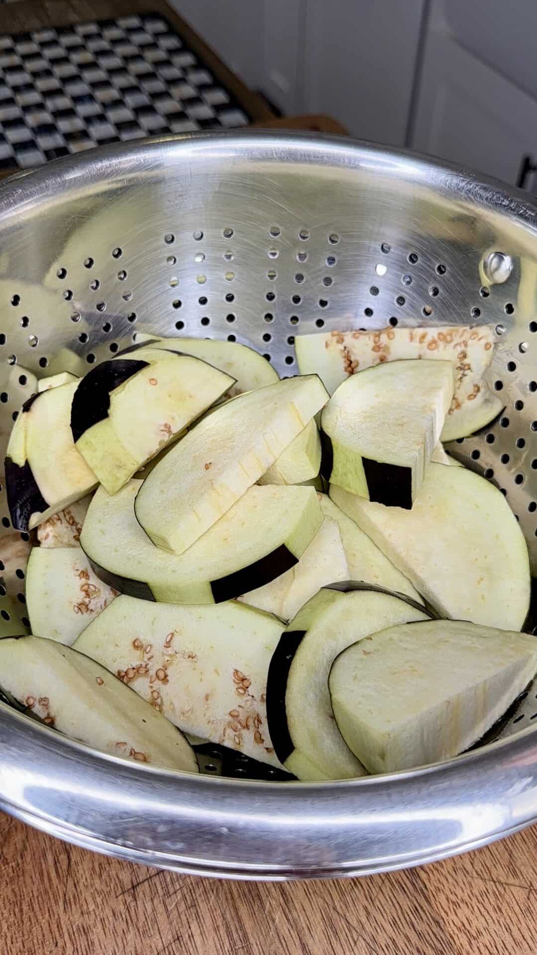 Sliced raw eggplant pieces in a metal colander on a wooden surface, with part of a kitchen counter and checkered mat visible in the background.