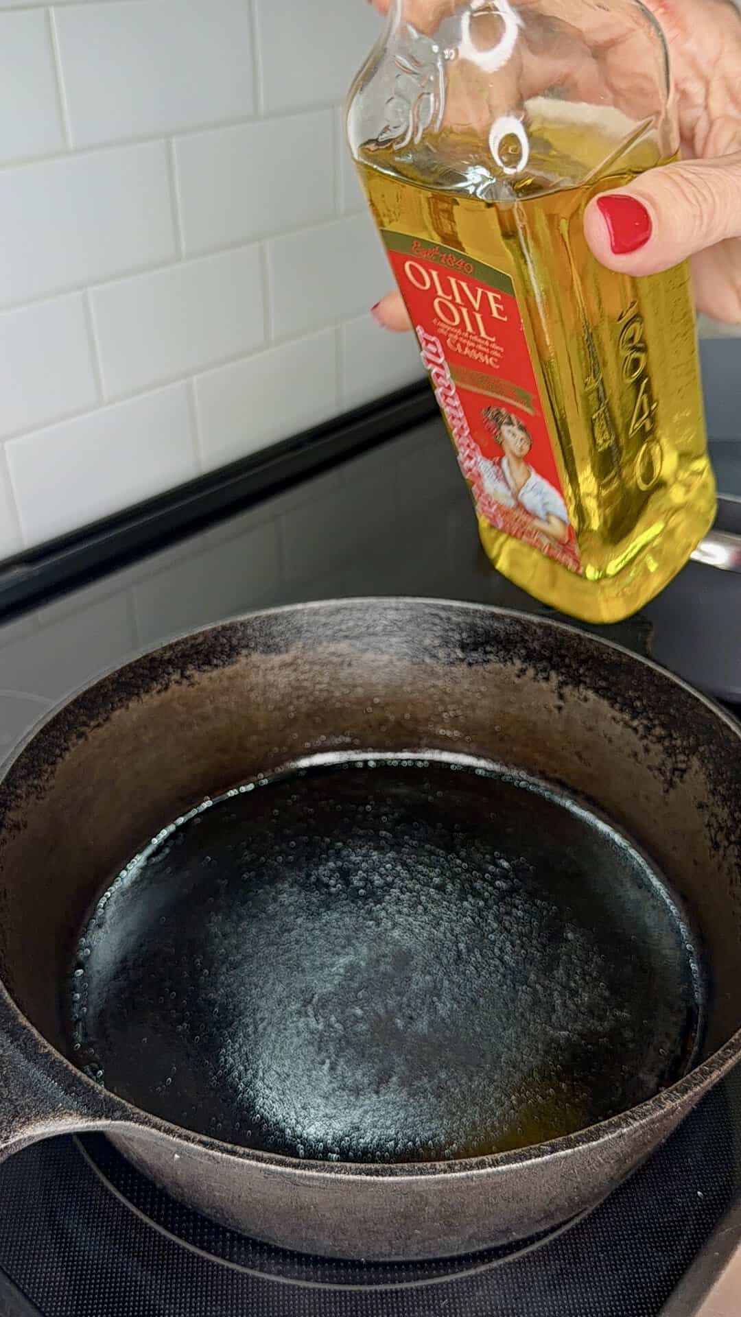 A hand with red nail polish holds a bottle of olive oil above a black cast iron skillet on a stovetop, preparing to pour oil into the pan. White tiled backsplash is visible in the background.