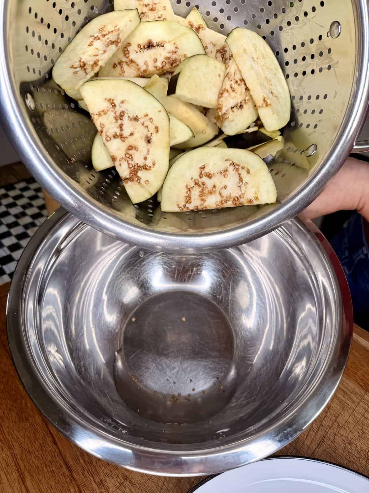 Sliced eggplant in a metal colander above a large empty metal bowl, both held over a wooden countertop.