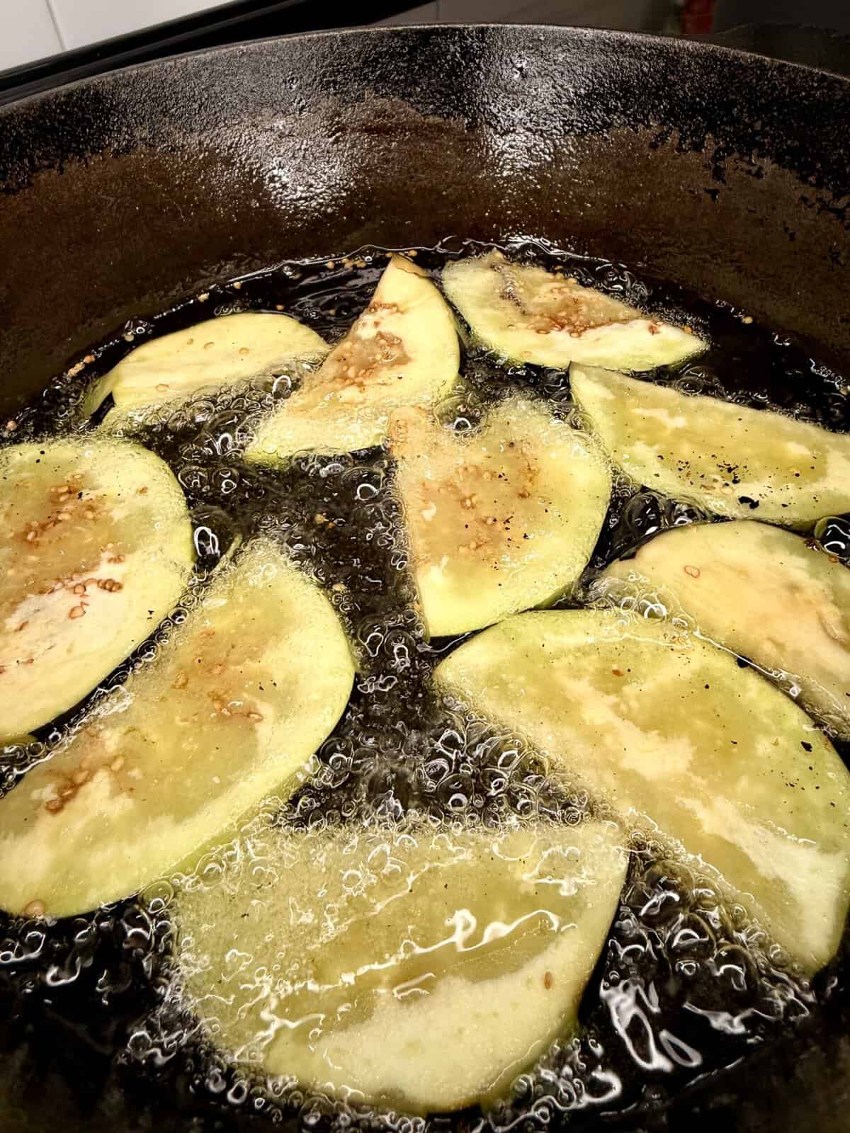 Several slices of eggplant are frying in hot oil in a dark pan, with bubbles forming around the edges of the sizzling vegetables.