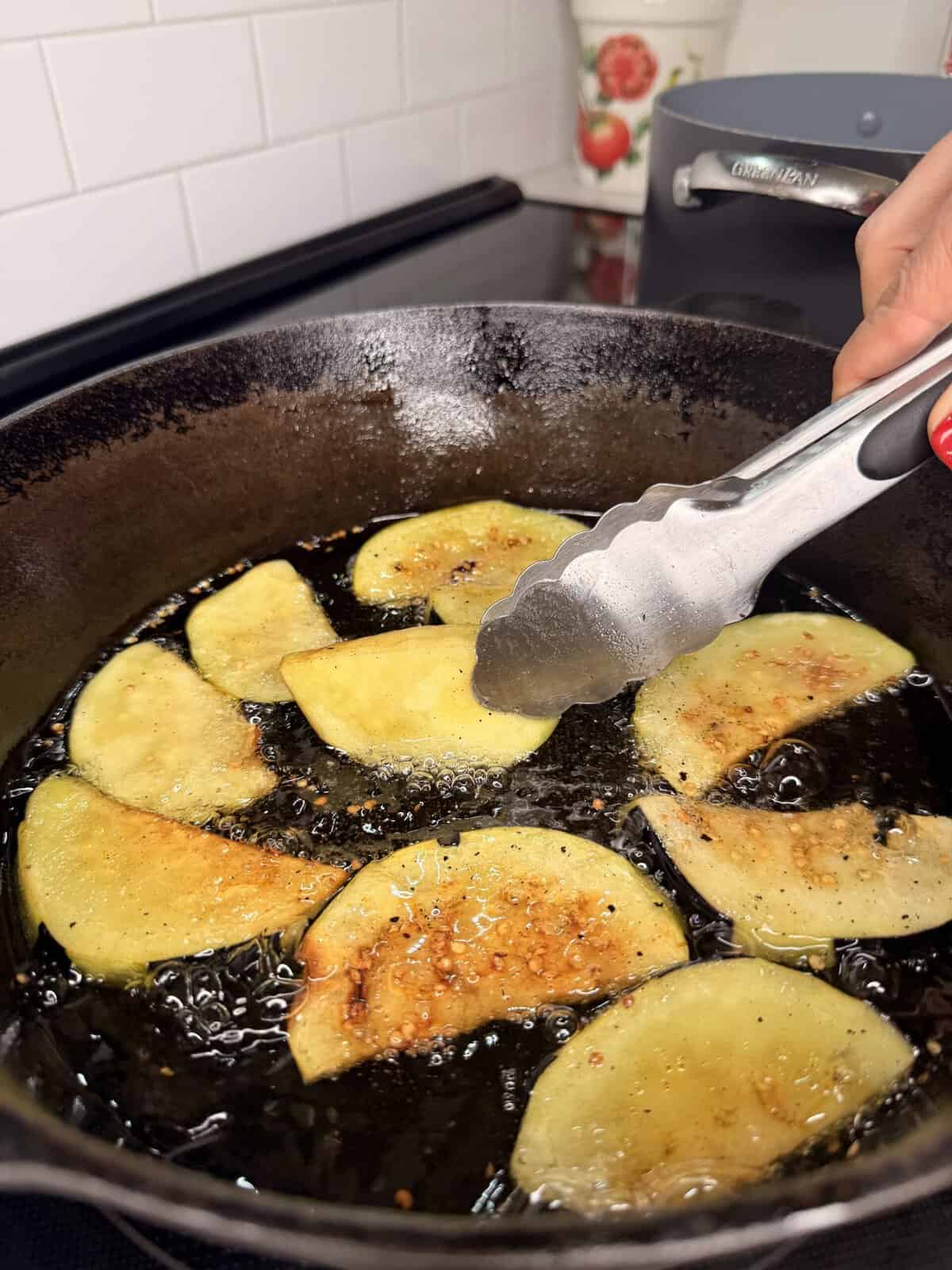 Sliced potatoes frying in oil in a cast iron pan, with a hand using metal tongs to turn a piece. White tile backsplash and a pot are visible in the background.