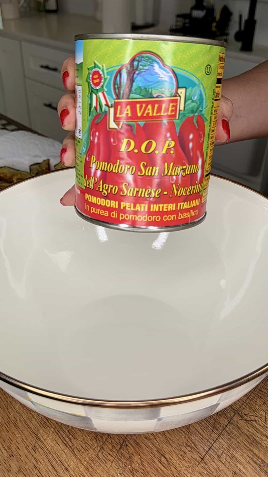 A hand with red nails holds a can of La Valle D.O.P. San Marzano tomatoes above a white bowl in a kitchen.