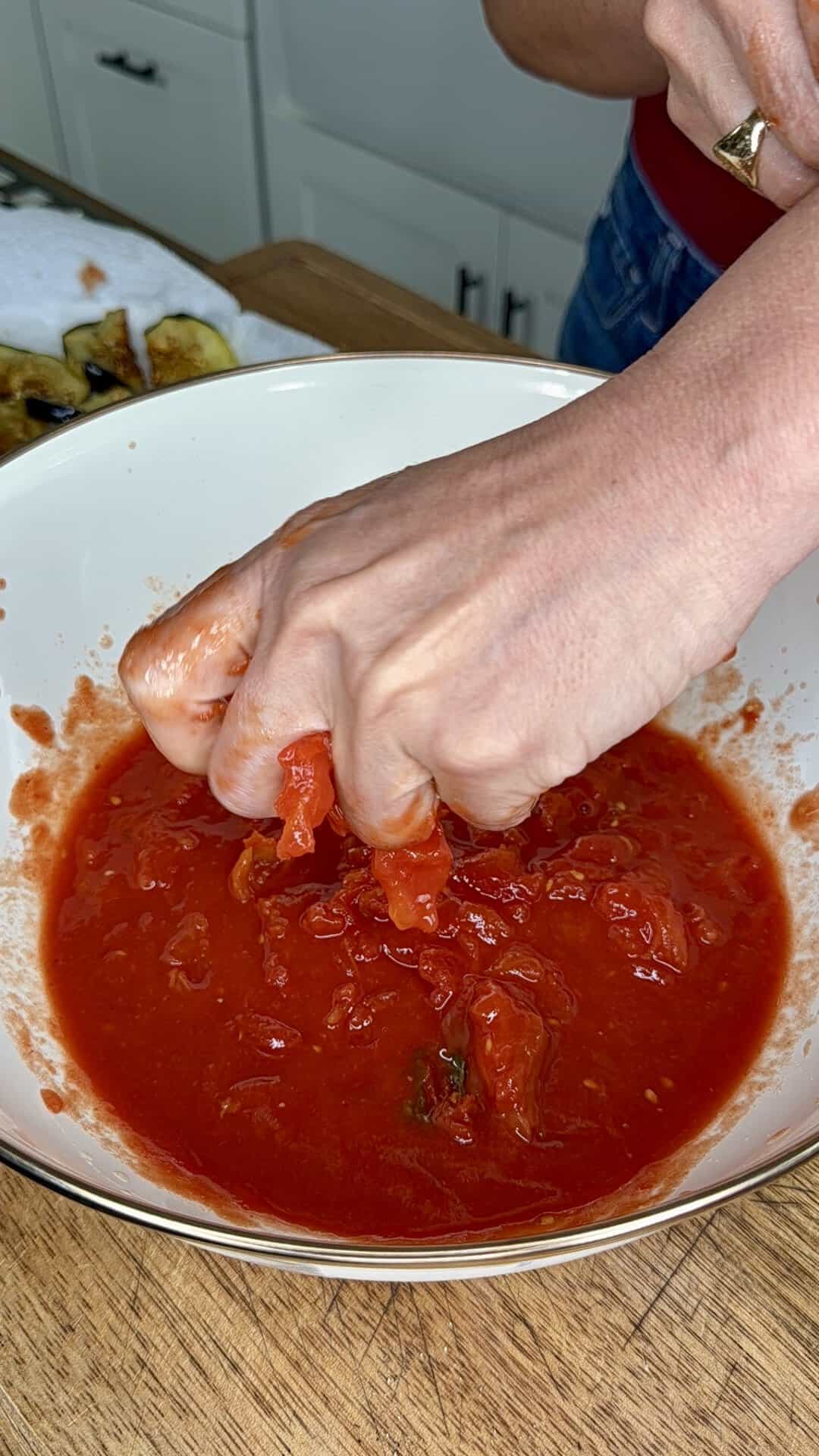 A person’s hand squeezes whole tomatoes in a white bowl, crushing them into a chunky tomato sauce. A cutting board with sliced vegetables is visible in the background on a wooden counter.