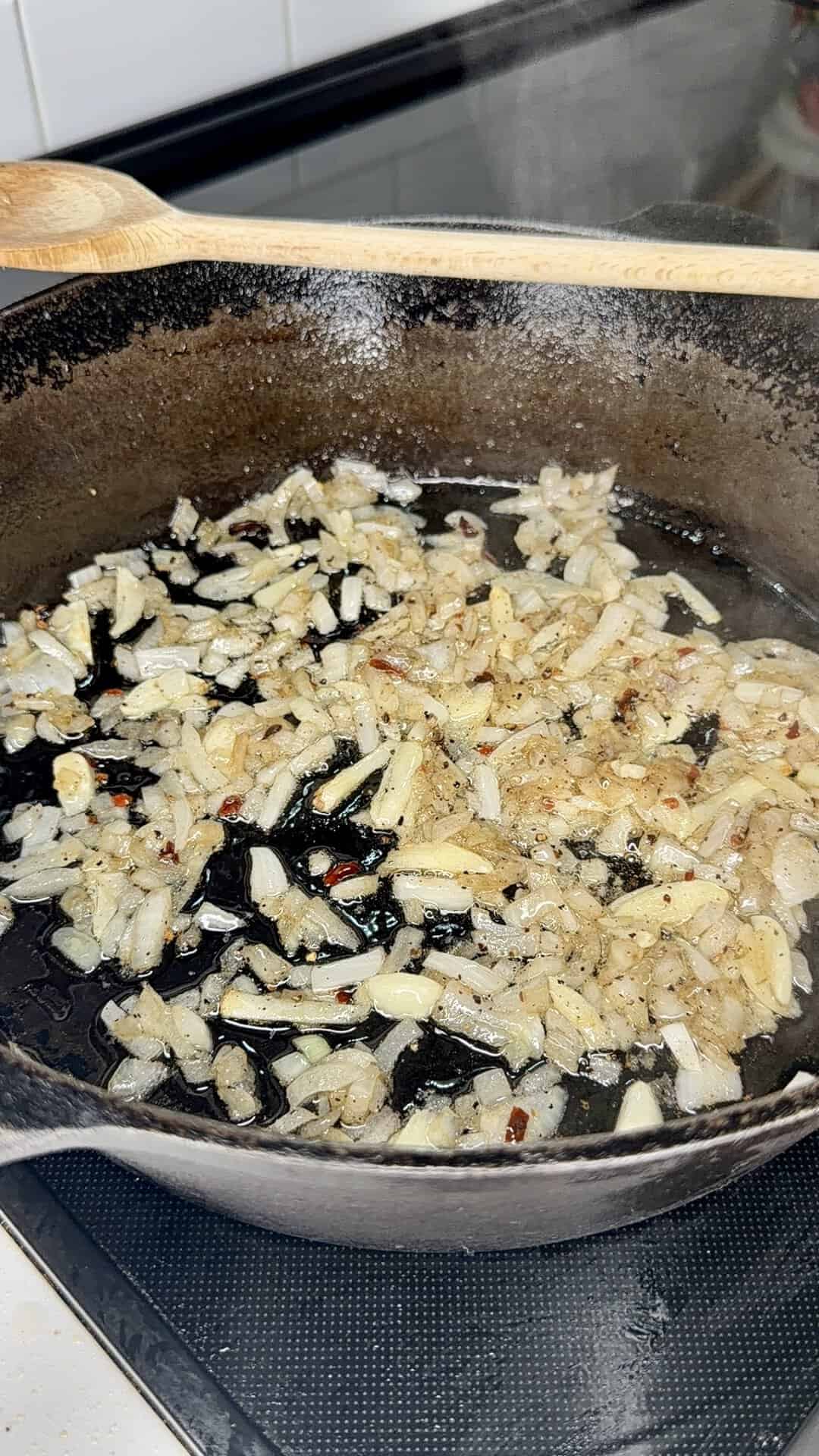 Diced onions sautéing in a black cast iron skillet with oil and seasonings on a stovetop, releasing steam as they cook.