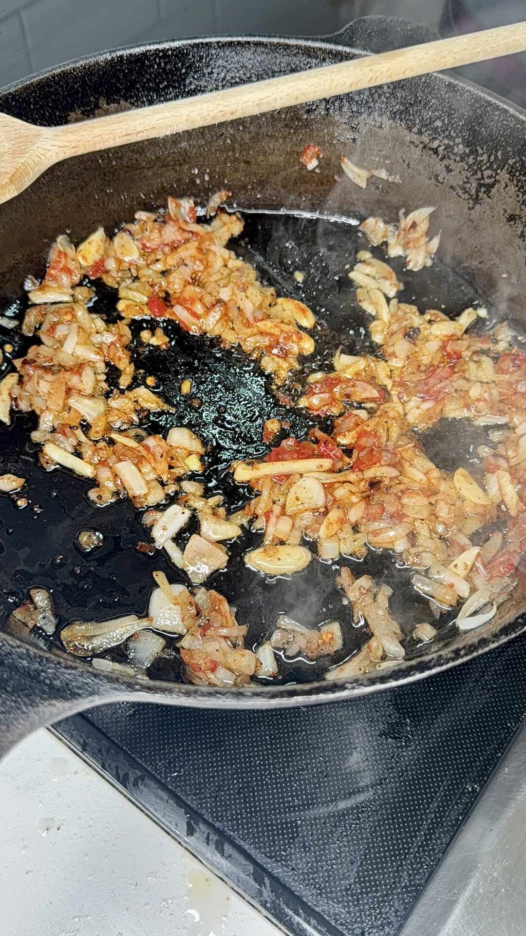 Chopped onions and diced tomatoes are sautéing in a cast iron skillet, with a wooden spoon resting on the edge. The mixture is starting to brown and caramelize.