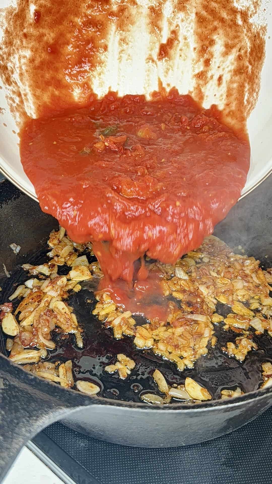 Tomato sauce is being poured from a white pot into a black skillet filled with sautéed onions and garlic. Steam rises from the pan as the sauce hits the hot ingredients.