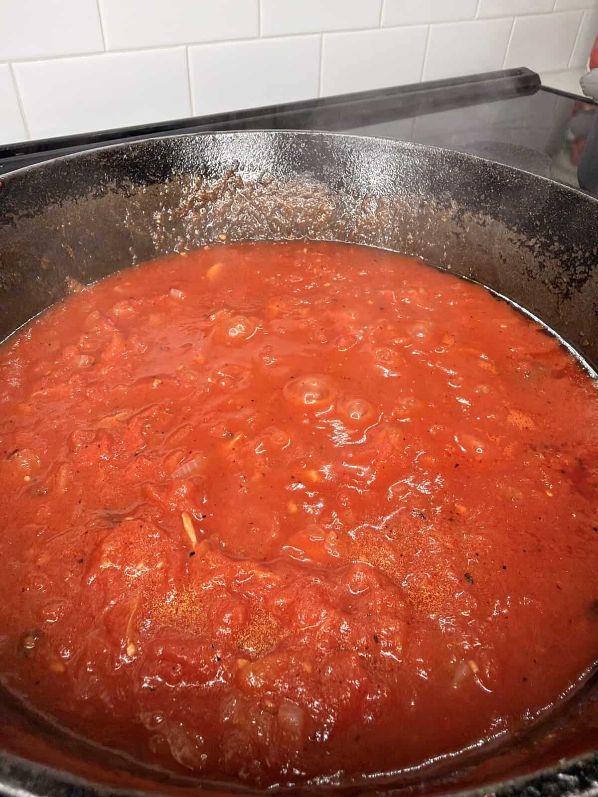A close-up of a simmering red tomato sauce with visible chunks of tomatoes and onions in a black cast iron pot on a stovetop.