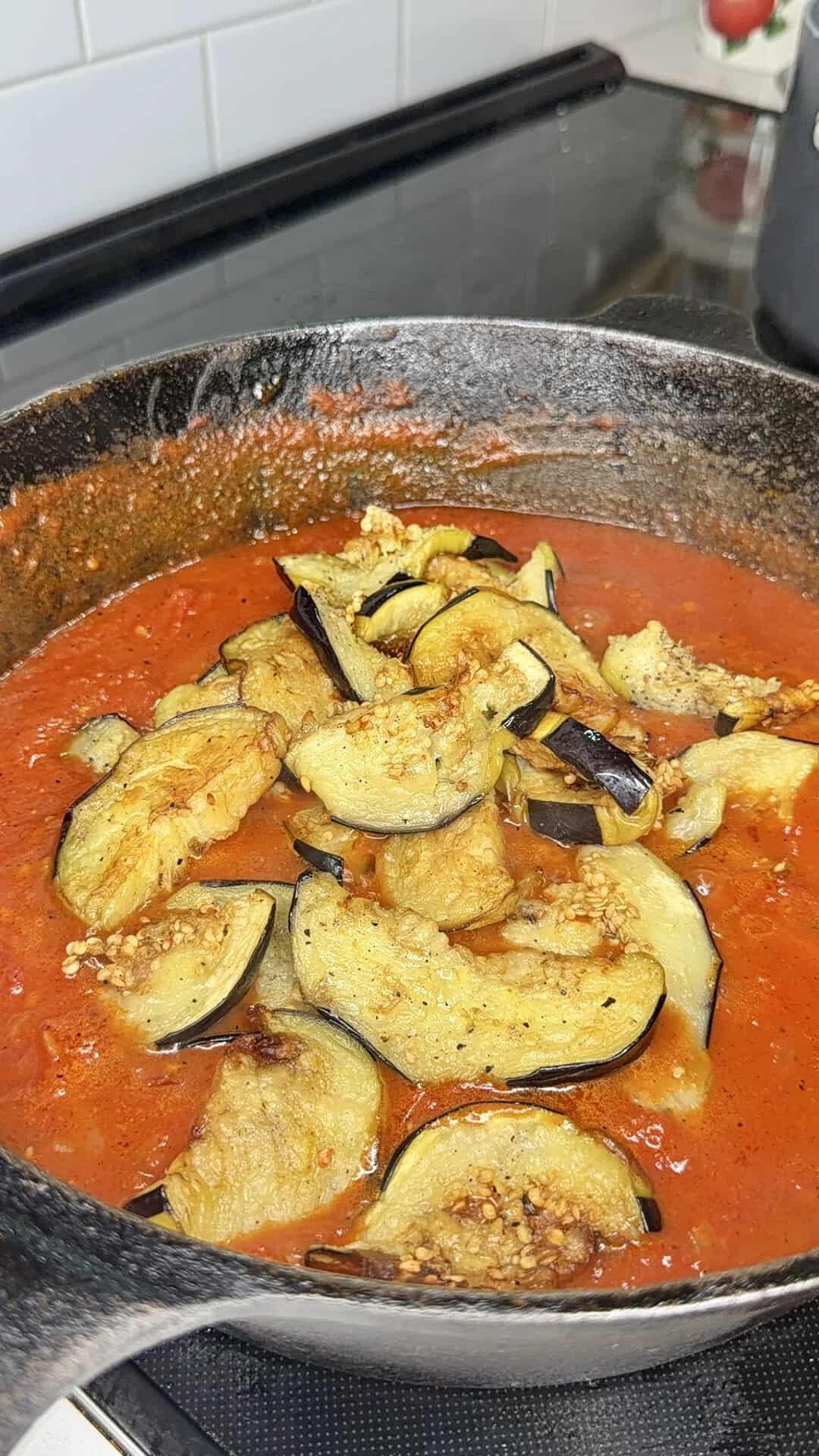 Sliced eggplant cooking in a skillet with red tomato sauce and minced garlic on a stovetop, surrounded by a black pan and white tiled backsplash.