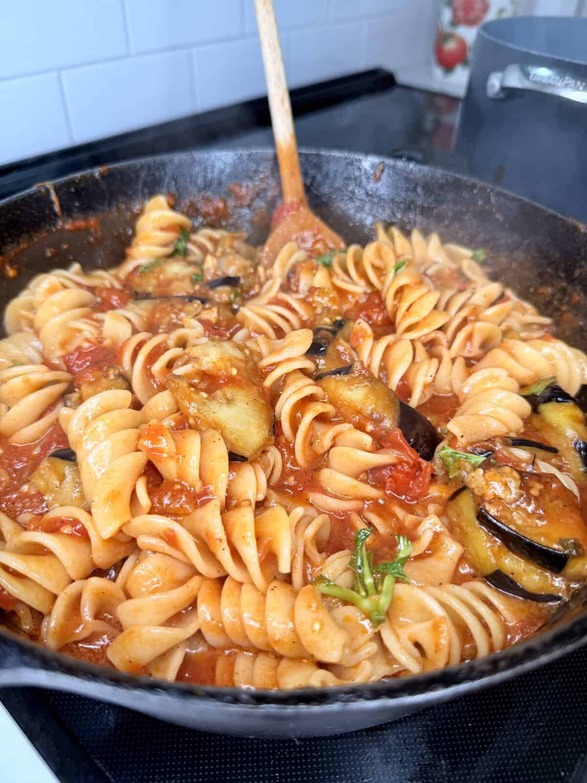 Rotini pasta with tomato sauce, eggplant slices, and fresh basil cooking in a black skillet, stirred with a wooden spoon on a stovetop.