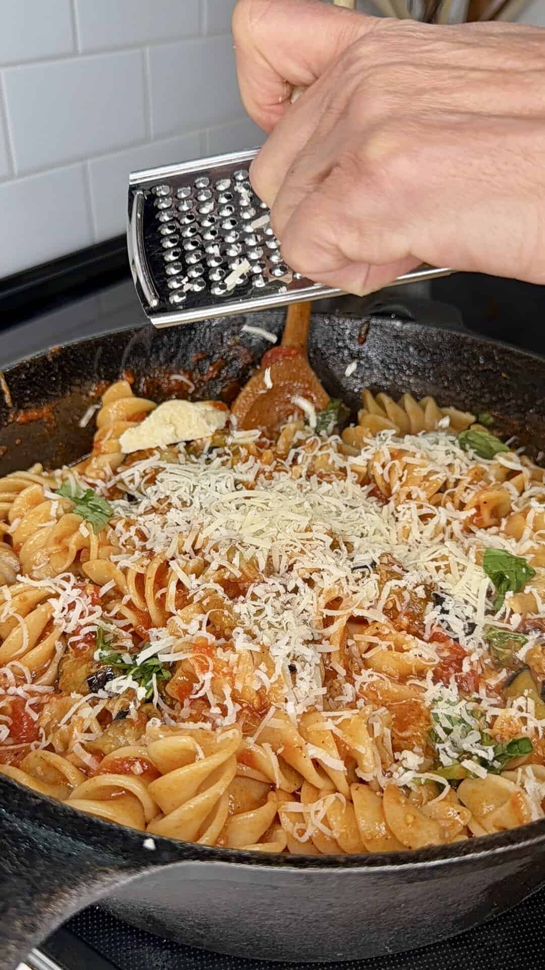 A hand grates cheese over a skillet filled with rotini pasta in tomato sauce, garnished with herbs, on a stove by a white tiled wall.