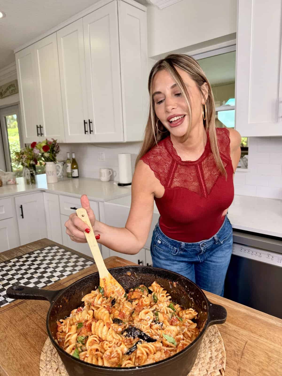 Woman in a red sleeveless top smiles while stirring pasta in a cast iron pan on a wooden counter in a bright, modern kitchen.