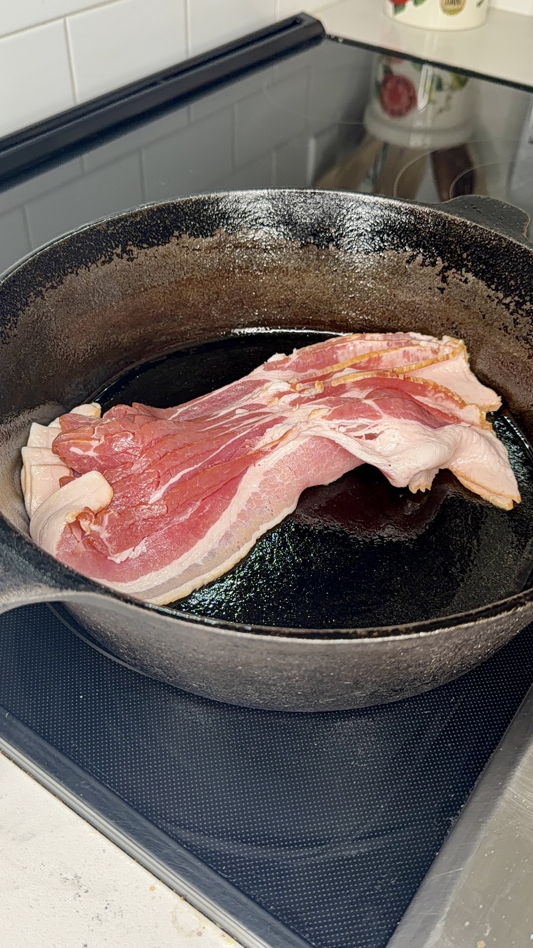 Two raw strips of bacon cooking in a black cast iron skillet on a stovetop, with a small amount of oil visible in the pan.