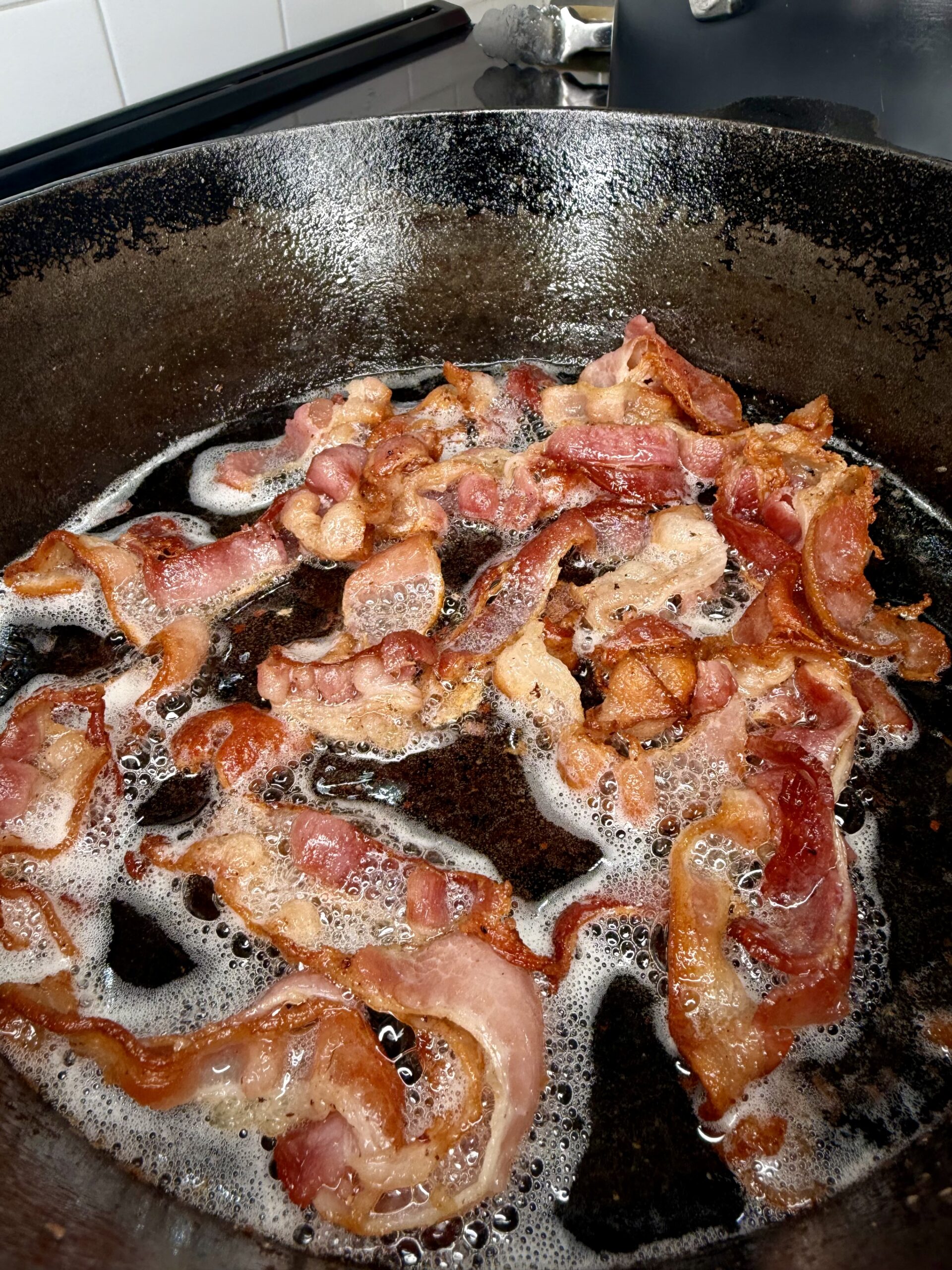Crispy bacon strips cooking in a black cast iron skillet with bubbling grease and foam around the edges.