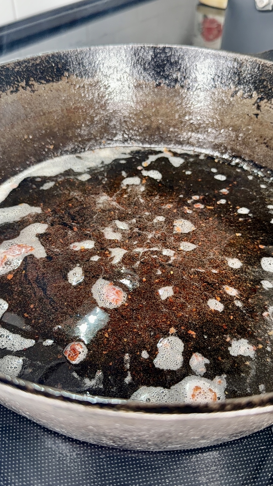 A close-up of a cast iron skillet on a stovetop with oil, bubbles, and bits of food residue covering the surface.