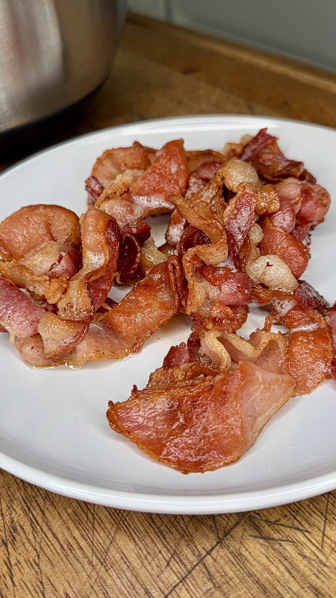 Crispy, cooked bacon strips are piled on a white plate placed on a wooden surface.