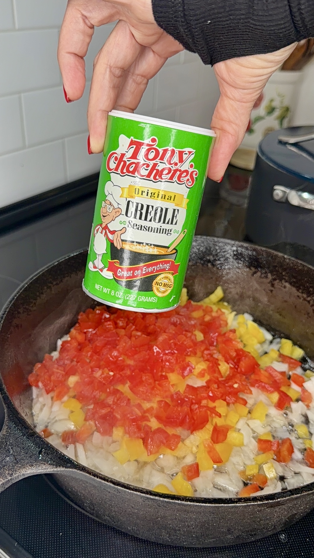 A hand holds Tony Chachere’s Creole Seasoning above a skillet with chopped onions, red, and yellow peppers sautéing on a stovetop.