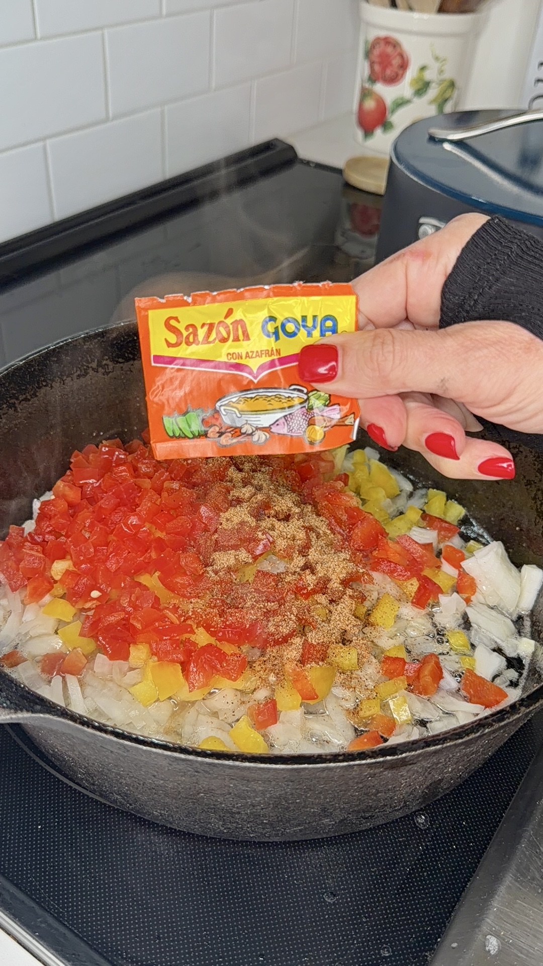 A hand sprinkles Sazon Goya seasoning onto chopped onions and red and yellow peppers cooking in a black skillet on a stove.