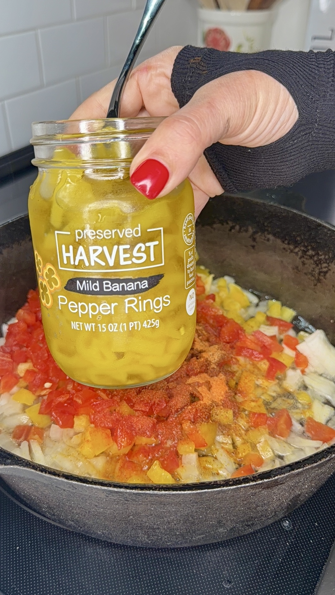 A hand with red nail polish holds a jar of mild banana pepper rings over a skillet filled with chopped vegetables, including red and yellow peppers and onions, on a stove.