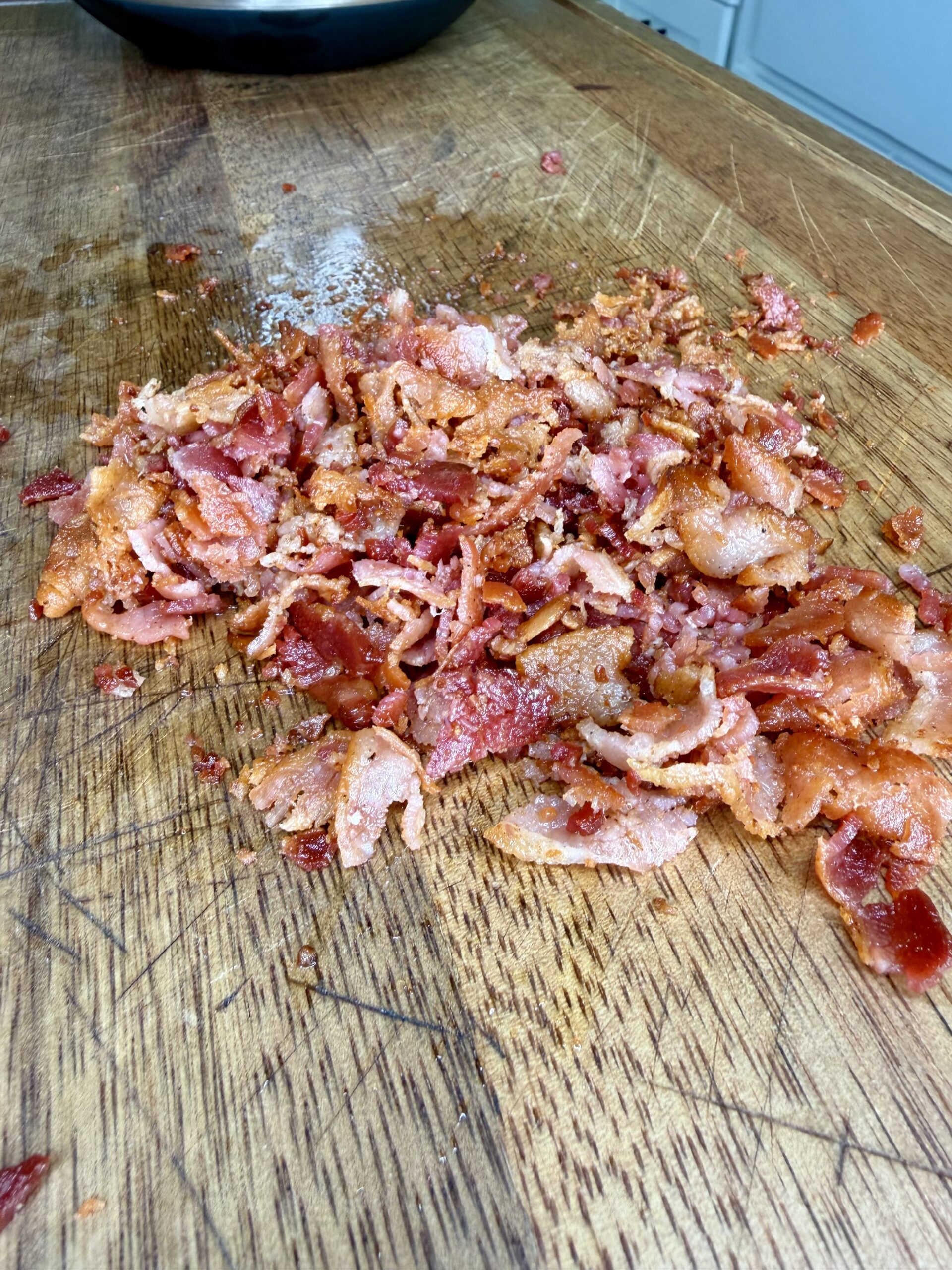 A pile of freshly cooked, crumbled bacon pieces sits on a wooden cutting board in a kitchen setting.