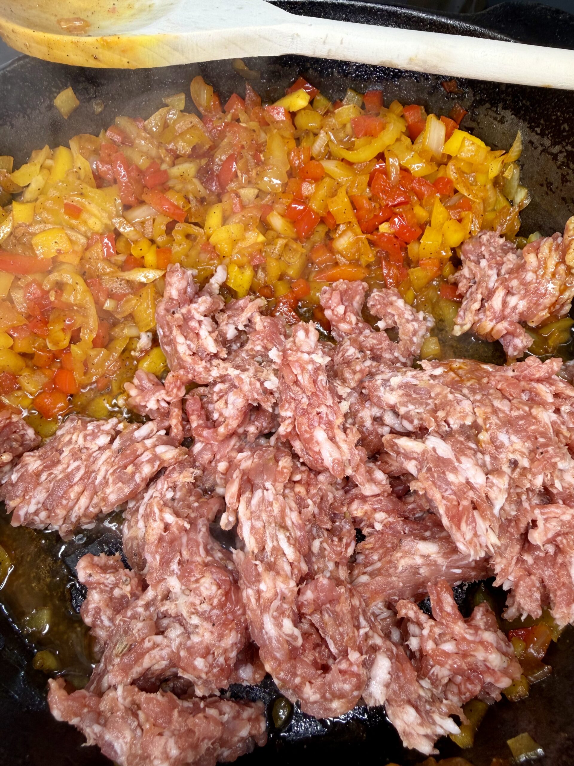 Ground meat cooking in a skillet with diced yellow and red bell peppers and onions. A wooden spoon rests at the top edge of the pan. The ingredients are sizzling and partially mixed together.