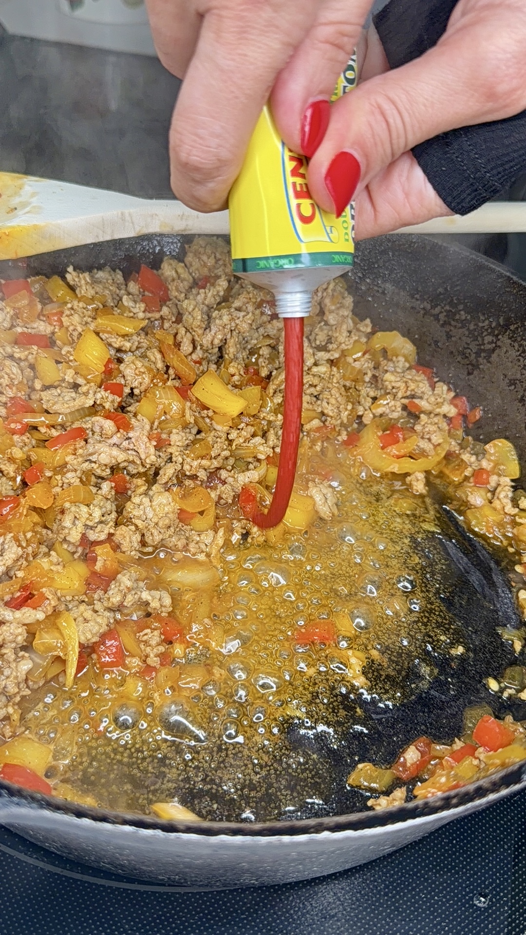 A hand with red nails squeezes tomato paste from a tube into a skillet of sizzling ground meat, onions, and red and yellow peppers.