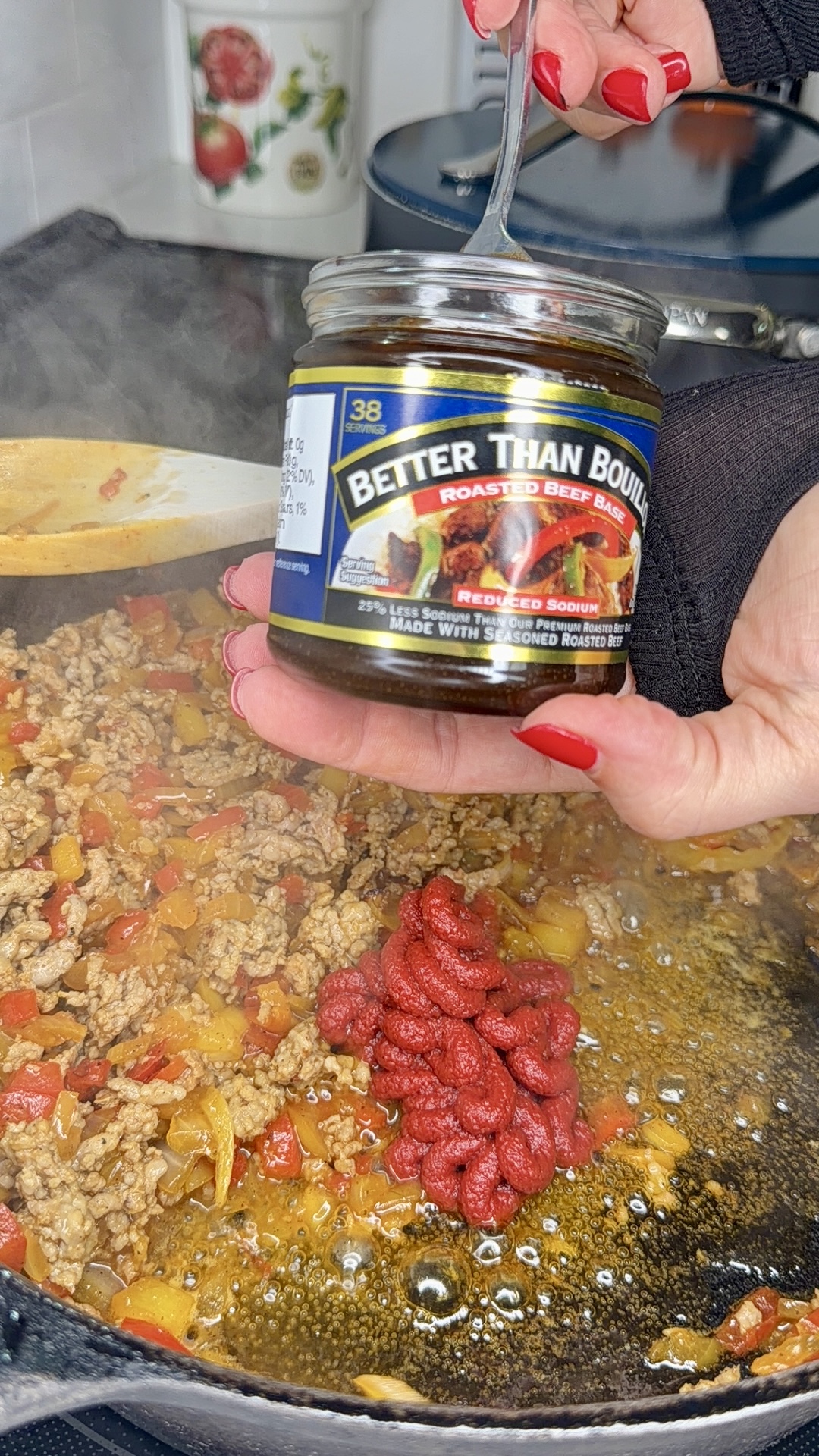 A hand holds a jar of Better Than Bouillon over a skillet with cooked ground meat, onions, and red peppers; tomato paste is added to the pan, and steam is rising from the food.