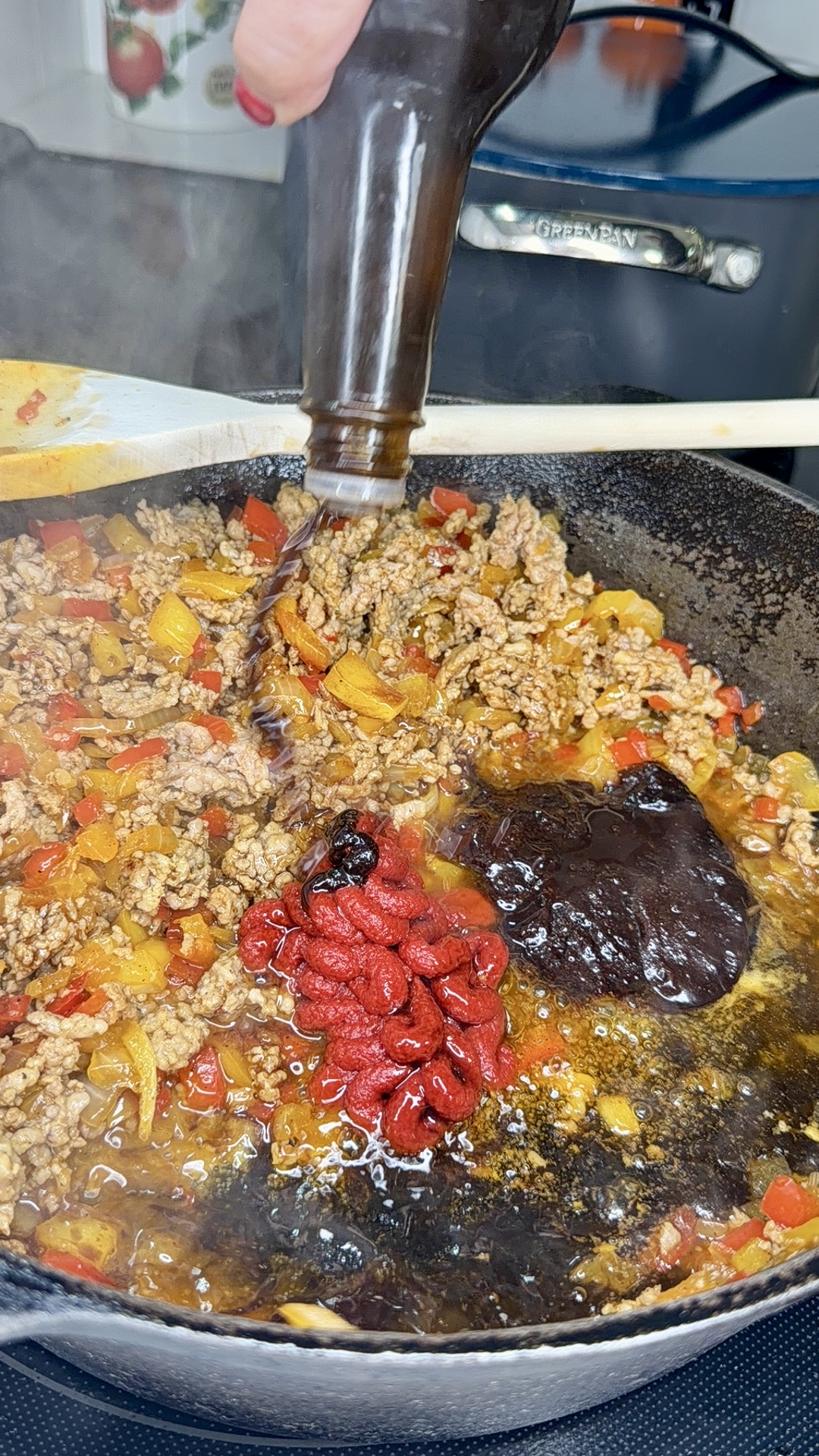 Ground meat, chopped peppers, tomato paste, and a dark sauce cook in a skillet. A hand pours liquid from a bottle into the pan, with wooden utensils and a lid visible in the background. Steam rises from the mixture.