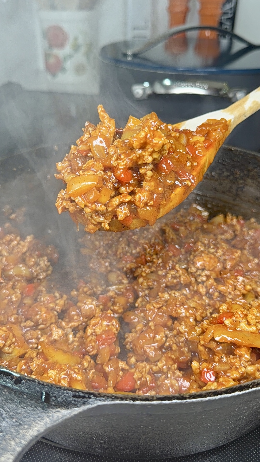 A steaming wooden spoon lifts a hearty serving of ground meat and vegetables in red sauce from a black cast iron pan on a stovetop.